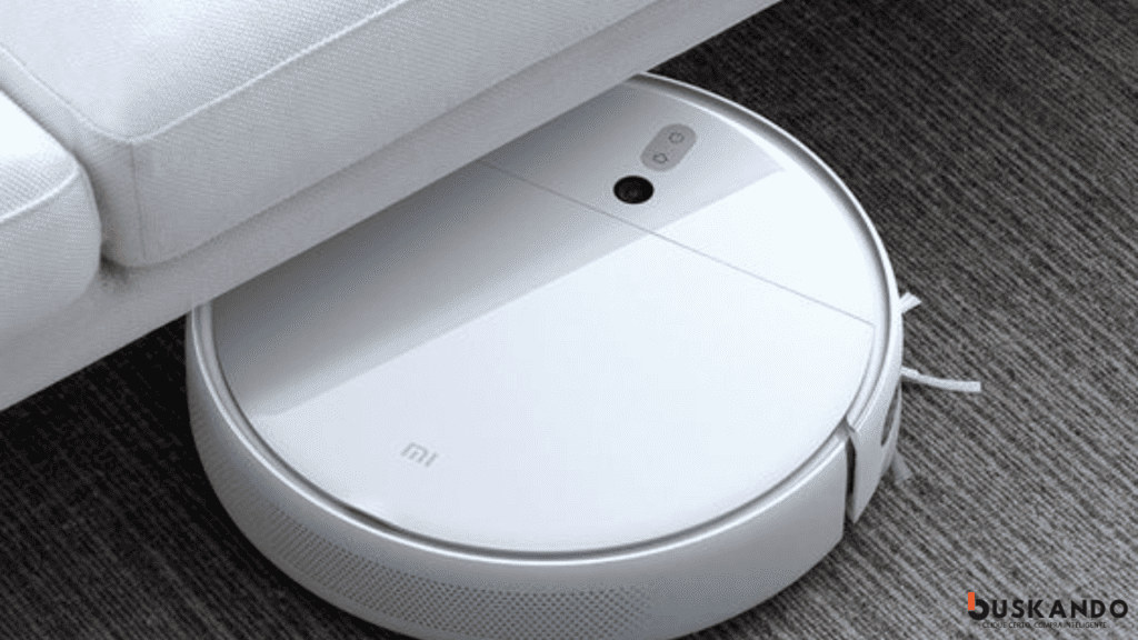 Robô aspirador de pó inteligente da marca Xiaomi navegando sob um sofá cinza, demonstrando sua capacidade de limpeza em espaços apertados.